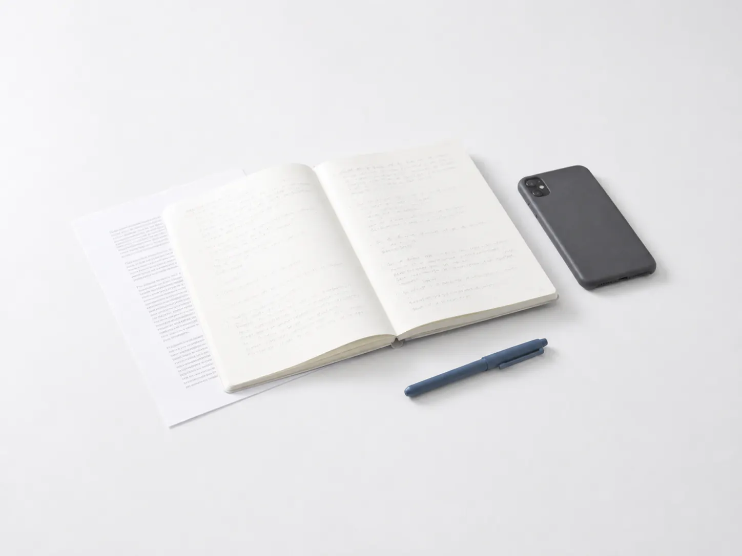An open notebook of handwriting beside a printed page, a dark phone, and a blue pen on a pale desk.